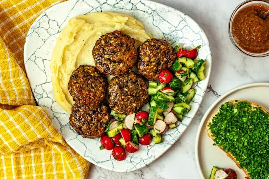 Five vegetarian Frikadellen meatballs on a bed of mashed potatoes with chopped vegetables served on a white plate with a side dip and bread.