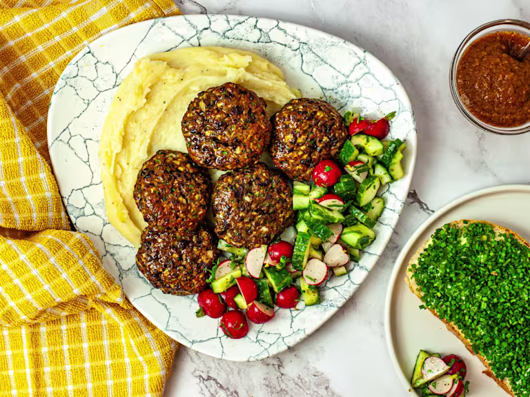 Five vegetarian Frikadellen meatballs on a bed of mashed potatoes with chopped vegetables served on a white plate with a side dip and bread.
