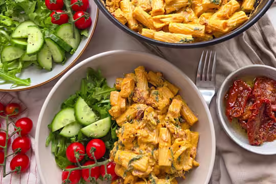 Vegetarian Tuscan Chicken Pasta in a white bowl with cucumber, sundried tomatoes and spinach on a white table.