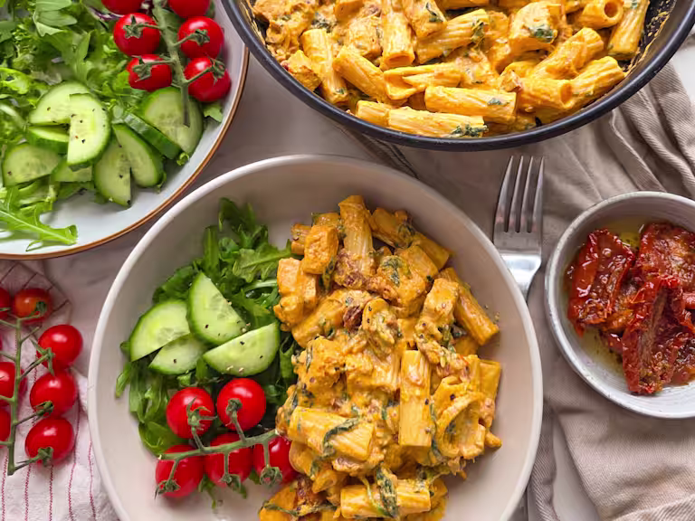 Vegetarian Tuscan Chicken Pasta in a white bowl with cucumber, sundried tomatoes and spinach on a white table.