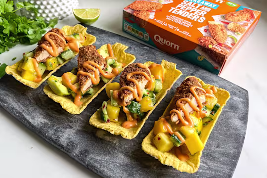 Four vegetarian Asian-style tacos with Quorn Salt & Pepper Tenders, topped with mango and cucumber salsa and an orange sauce. Served on a dark serving tray with fresh coriander and lime in the background.