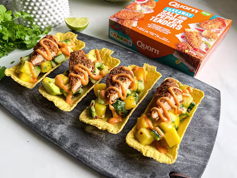 Four vegetarian Asian-style tacos with Quorn Salt & Pepper Tenders, topped with mango and cucumber salsa and an orange sauce. Served on a dark serving tray with fresh coriander and lime in the background.