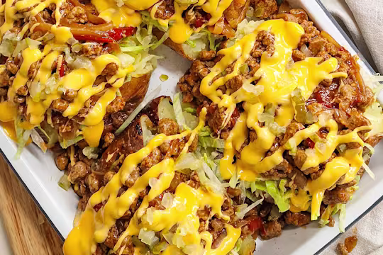 Cheeseburger Jacket Potatoes with Quorn Mince, topped with melted cheese, onion, cherry tomatoes, garlic, paprika, lettuce and gherkins on a white plate on a wooden board.
