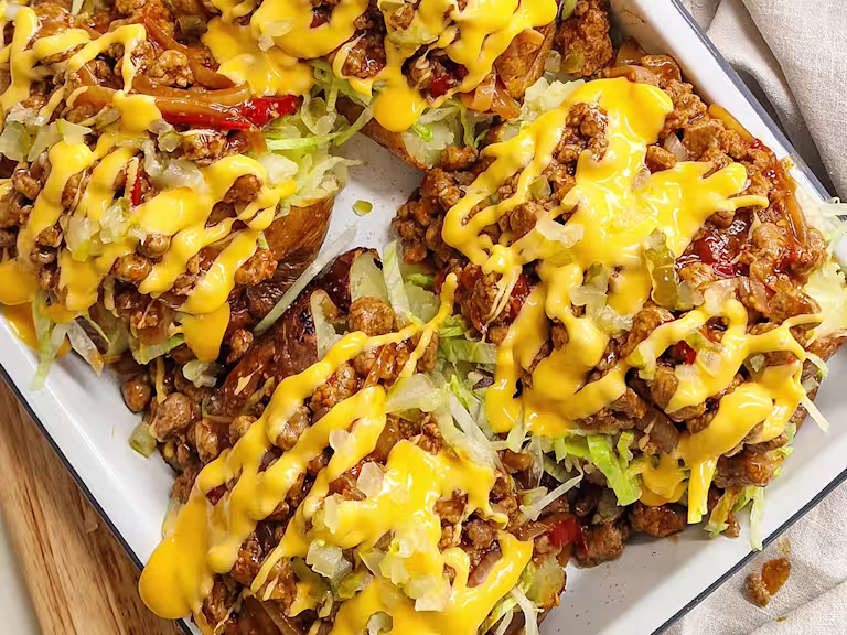 Cheeseburger Jacket Potatoes with Quorn Mince, topped with melted cheese, onion, cherry tomatoes, garlic, paprika, lettuce and gherkins on a white plate on a wooden board.