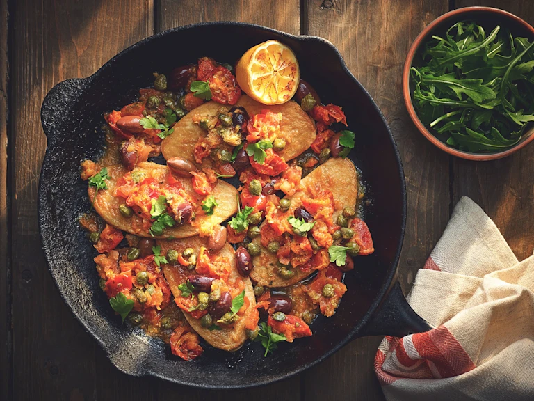 Quorn Fillets topped with a sauce made of kalamata olives, capers, and tomatoes topped with parsley and half of a charred lemon in a cast iron skillet with a terra cotta bowl of arugula on the side.