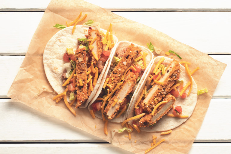Three tacos in flour tortillas filled with sliced Quorn Vegan Meatless Chipotles Cutlets, cheese, refried beans, lettuce, and pico de gallo salsa on a piece of brown paper.