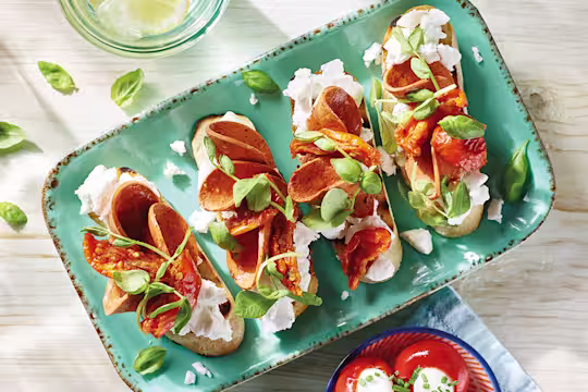 Four servings of vegan bruschetta topped with Violife Greek White Block, Quorn Vegan Pepperoni, sunblush tomatoes, and basil on a pale turquoise tray.