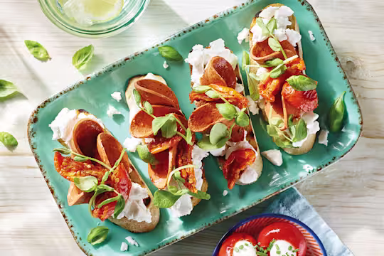 Four servings of vegan bruschetta topped with Violife Greek White Block, Quorn Vegan Pepperoni, sunblush tomatoes, and basil on a pale turquoise tray.