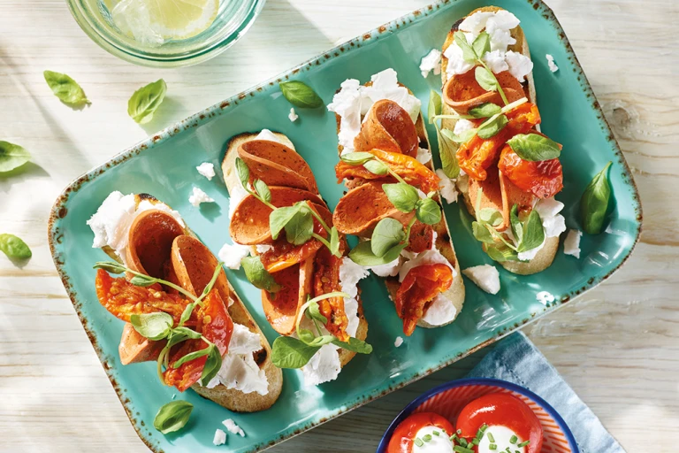 Four servings of vegan bruschetta topped with Violife Greek White Block, Quorn Vegan Pepperoni, sunblush tomatoes, and basil on a pale turquoise tray.