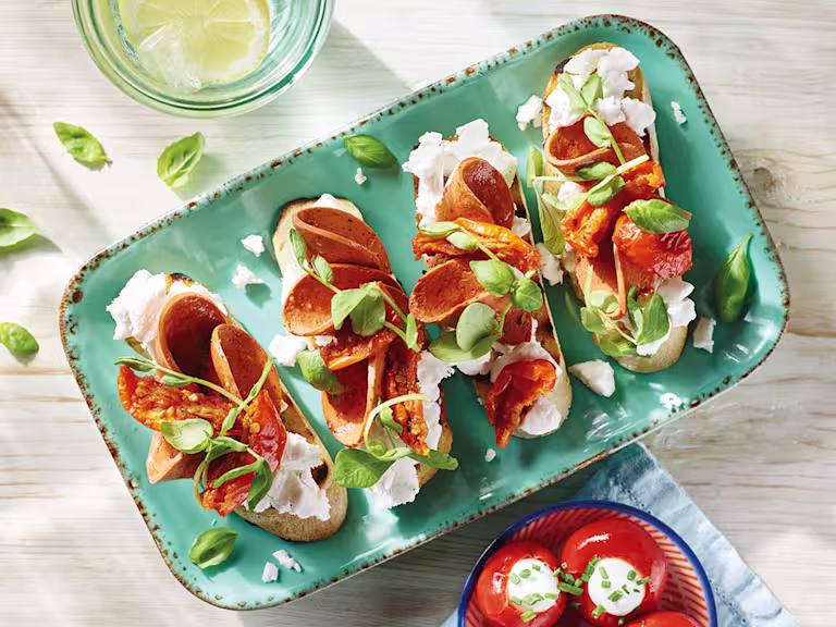 Four servings of vegan bruschetta topped with Violife Greek White Block, Quorn Vegan Pepperoni, sunblush tomatoes, and basil on a pale turquoise tray.