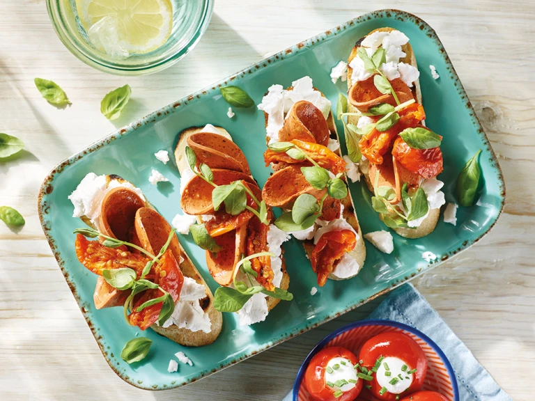 Four servings of vegan bruschetta topped with Violife Greek White Block, Quorn Vegan Pepperoni, sunblush tomatoes, and basil on a pale turquoise tray.