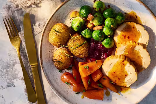 A Quorn Roast with Spiced Hot Honey Sauce, carrots, brussels sprouts and hassleback potatoes on a white plate.
