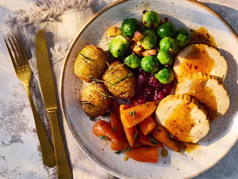 A Quorn Roast with Spiced Hot Honey Sauce, carrots, brussels sprouts and hassleback potatoes on a white plate.