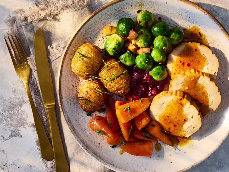 A Quorn Roast with Spiced Hot Honey Sauce, carrots, brussels sprouts and hassleback potatoes on a white plate.