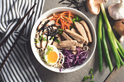 A bowl of ramen noodles topped with sliced mushrooms, edamame, shredded carrots, shredded purple cabbage, green onions, half of a half-cooked egg, nori, and Quorn Strips, with chopsticks and green onions on either side.