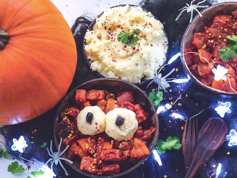 Pumpkin curry with Quorn Pieces in a bowl with balls of mash on top for eyes next to a pumpkin, bowl of mash and ghost lights