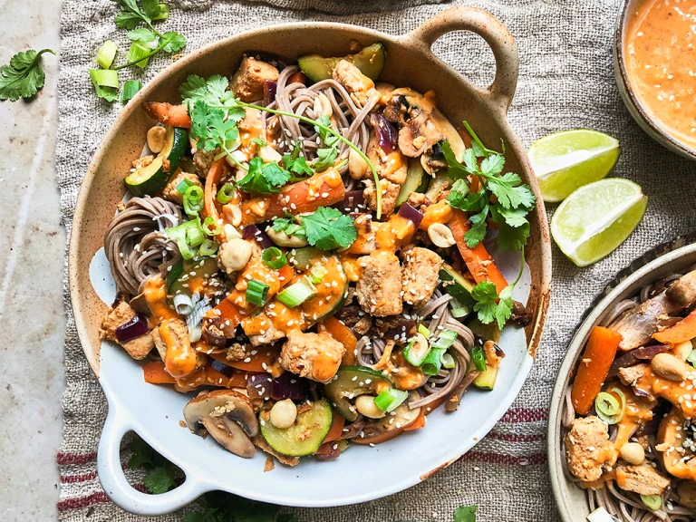 A bowl of vegan satay noodles topped with Quorn Vegan Pieces, carrots, mushrooms, courgette, green onions, peanuts, sesame seeds, coriander, and satay sauce.
