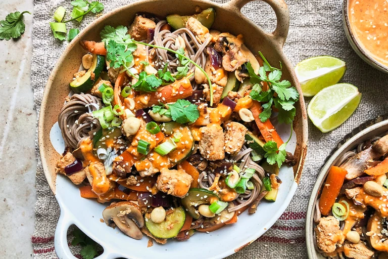 A bowl of vegan satay noodles topped with Quorn Vegan Pieces, carrots, mushrooms, courgette, green onions, peanuts, sesame seeds, coriander, and satay sauce.