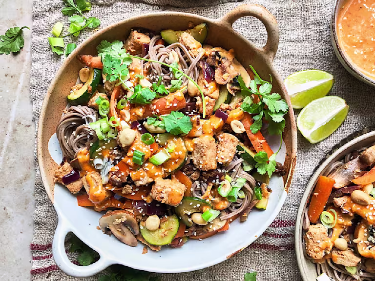 A bowl of vegan satay noodles topped with Quorn Vegan Pieces, carrots, mushrooms, courgette, green onions, peanuts, sesame seeds, coriander, and satay sauce.