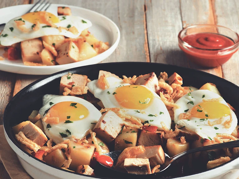 A skillet full of potatoes and cubed Quorn Meatless Roast topped with three fried eggs and crispy onions.