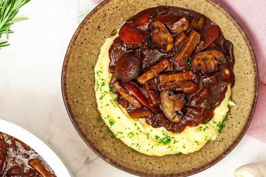 Vegetarian Beef Bourguignon with Quorn Strips on a plate with mashed potatoes on a marble work surface with a pink cloth.