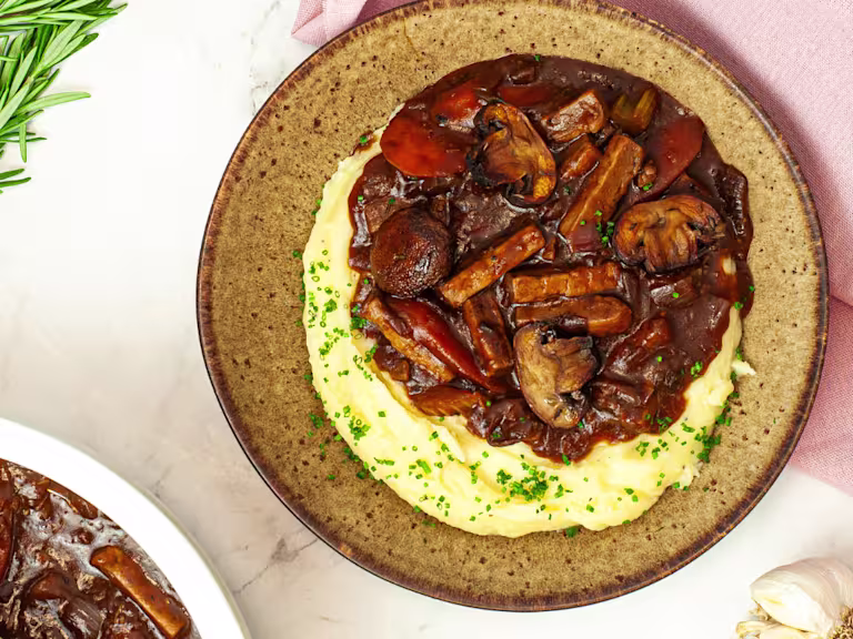 Vegetarian Beef Bourguignon with Quorn Strips on a plate with mashed potatoes on a marble work surface with a pink cloth.
