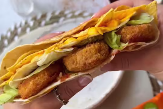 Hands holding a Quorn Cheesy Nacho Nuggs in a wrap wtih tortilla chips and salad