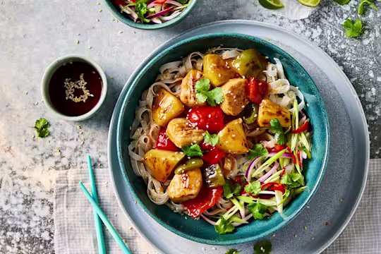 A bowl of Quorn vegetarian sweet and sour chicken with sesame noodles with Quorn Fillet Pieces visible on top with a side of sauce.
