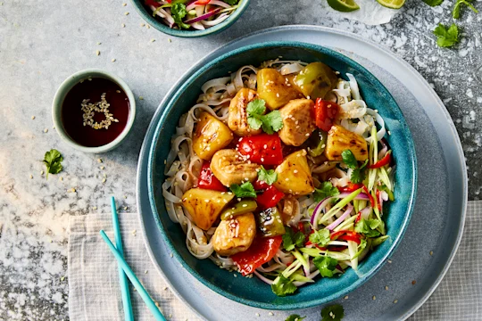 A bowl of Quorn vegetarian sweet and sour chicken with sesame noodles with Quorn Fillet Pieces visible on top with a side of sauce.
