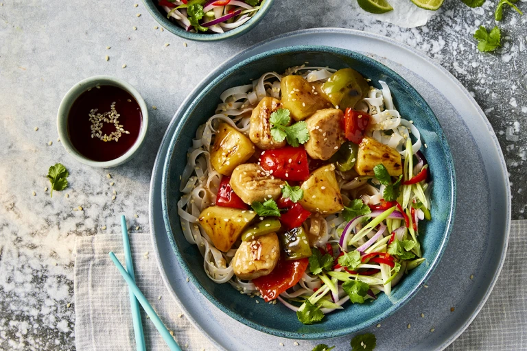 A bowl of Quorn vegetarian sweet and sour chicken with sesame noodles with Quorn Fillet Pieces visible on top with a side of sauce. 