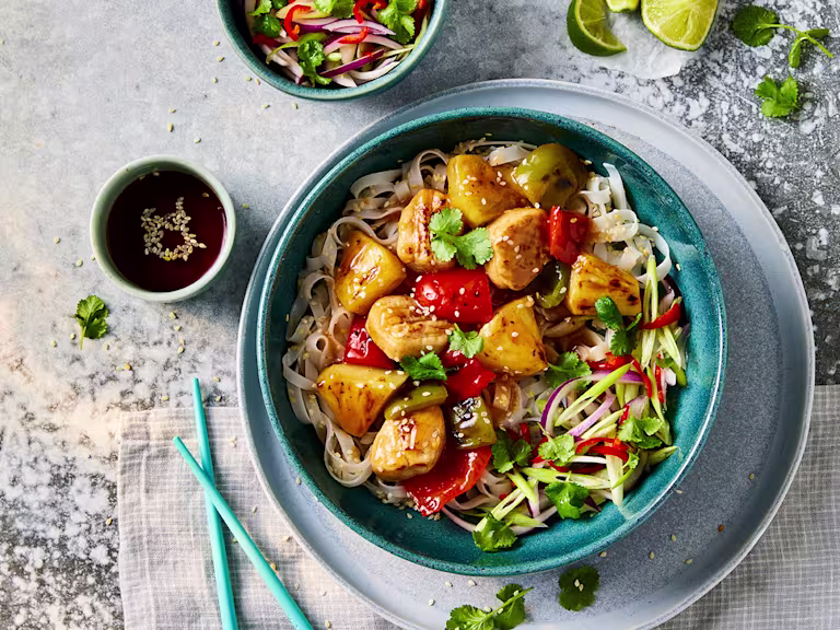 A bowl of Quorn vegetarian sweet and sour chicken with sesame noodles with Quorn Fillet Pieces visible on top with a side of sauce.