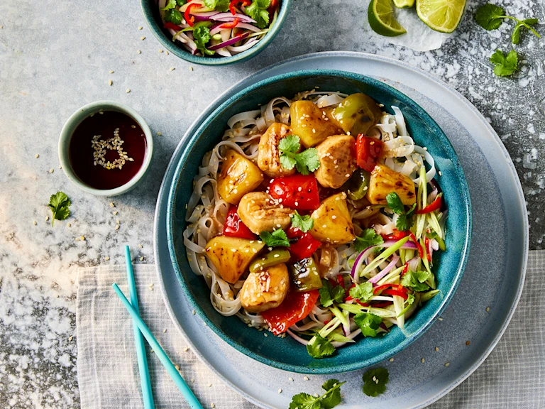 A bowl of Quorn vegetarian sweet and sour chicken with sesame noodles with Quorn Fillet Pieces visible on top with a side of sauce.