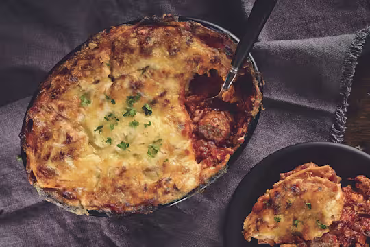 A lasagne made with Quorn Swedish Style Balls topped with cheese and parsley in a black baking dish.