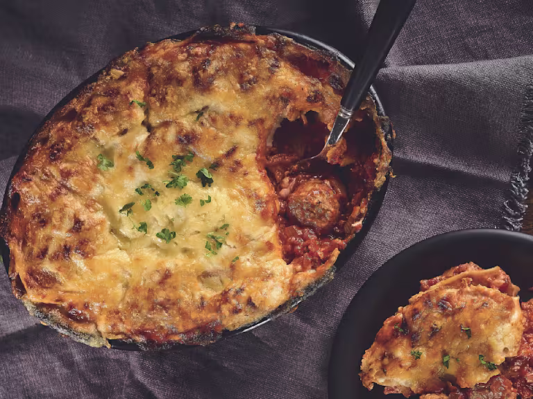 A lasagne made with Quorn Swedish Style Balls topped with cheese and parsley in a black baking dish.
