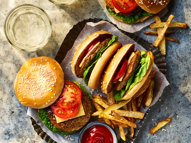 Four Quorn Classic Vegetarian Cheeseburger with one open to show the toppings served alongside fries and sauce. 