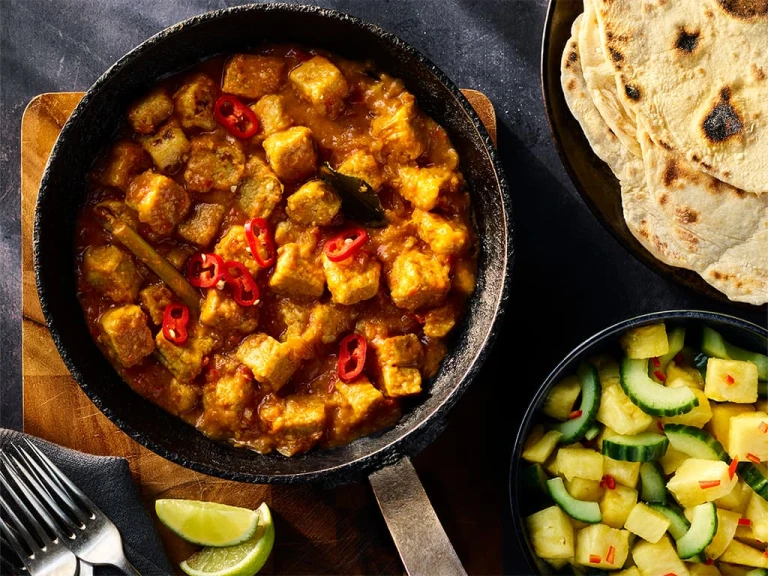 Quorn Vegetarian Rendang Curry served in a dark dish with a side of pineapple and cucumber salad and flatbread. 