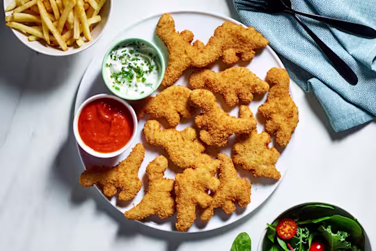 A plate of Quorn Roarsomes Vegan Dino Nuggets with homemade ketchup and vegan yogurt dip on the side as well as a bowl of chips and a bowl of salad.
