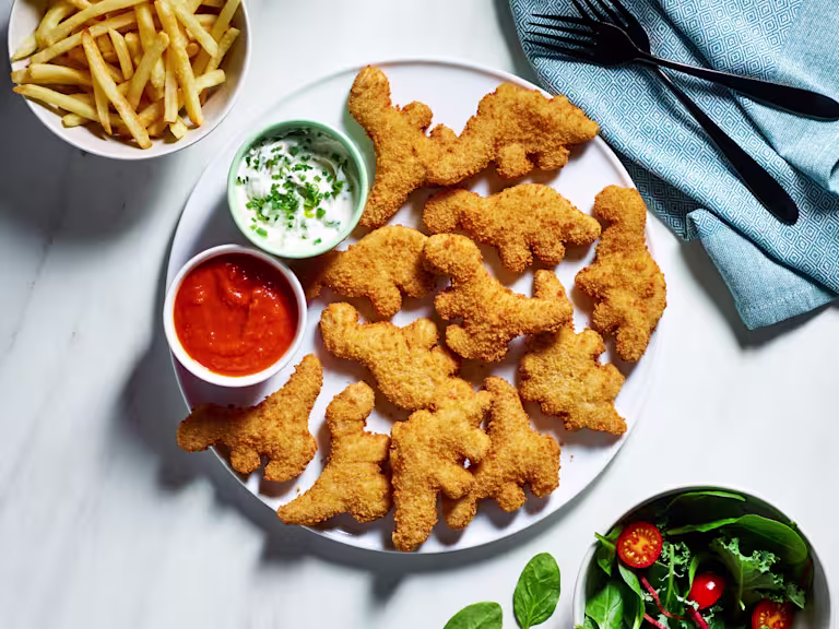 A plate of Quorn Roarsomes Vegan Dino Nuggets with homemade ketchup and vegan yogurt dip on the side as well as a bowl of chips and a bowl of salad.