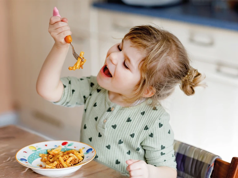 A young child with a bowl full of pasta with some on a fork smiling.