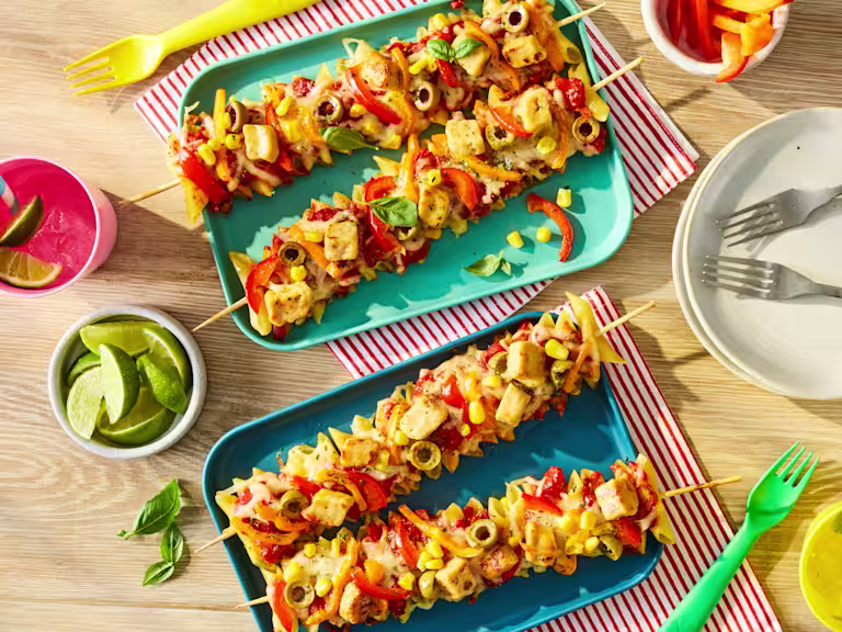 Penne pasta and Quorn Pieces threaded onto skewers with sliced peppers, topped with pizza sauce, melted mozzarella and olives. Served on a plate.