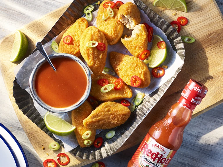 Vegan Buffalo wings topped with red and green chillies with the sauce on the side next to a bottle of Franks redhot original Cayenne Pepper Sauce.