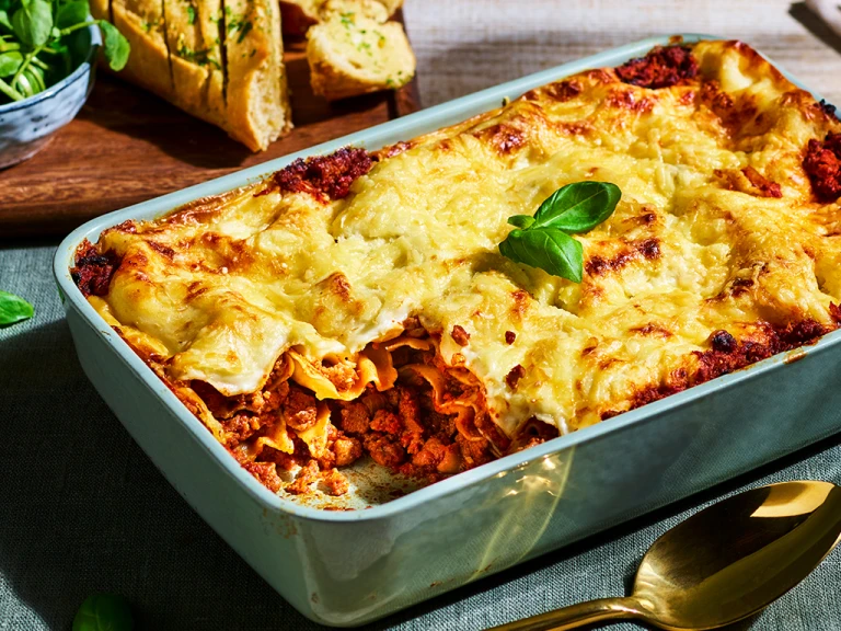 A white dish of vegetarian lasagna with Quorn Grounds with one portion already served to show the layers in the dish.
