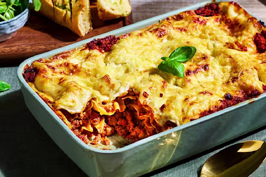 Vegetarian lasagne with Quorn Mince in a light blue baking dish with a corner slice cut, garlic bread in the background and basil garnish.