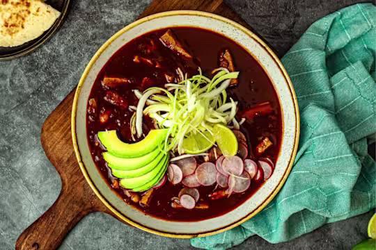 Pozole soup garnished with avocado slices, shredded cabbage served in a bowl on a wood board with a green napkin and side of bread.
