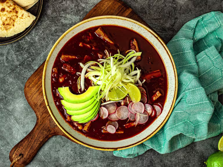 Pozole soup garnished with avocado slices, shredded cabbage served in a bowl on a wood board with a green napkin and side of bread.