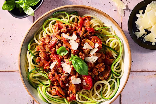 A courgette spaghetti Bolognese made with a Quorn Mince ragu in a white bowl on a kitchen worktop. Vegetarian parmesan and basil serving suggestions at the side.