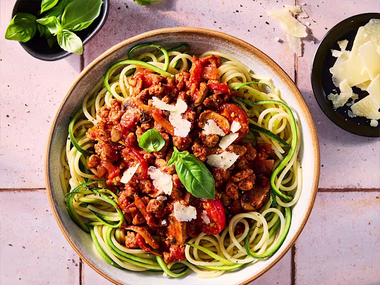 A courgette spaghetti Bolognese made with a Quorn Mince ragu in a white bowl on a kitchen worktop. Vegetarian parmesan and basil serving suggestions at the side.