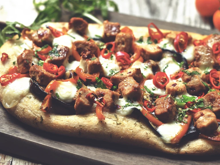 A flatbread pizza topped with cheese, eggplant, pepper, chile, and Quorn Pieces.