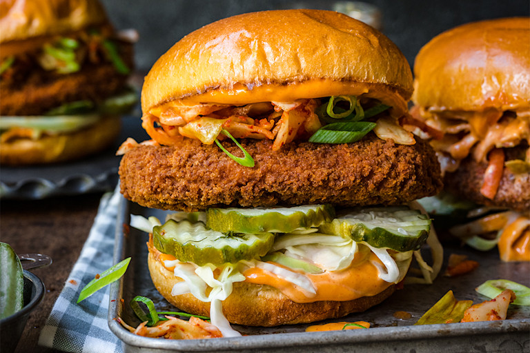 A sandwich filled with a Quorn Fillet, avocado, tomato slices, onion slices, and lettuce, drizzled with a sauce.