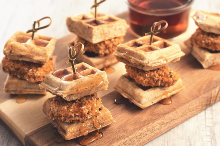 Five individual mini waffle sliders filled with Quorn Tex Mex Nuggets each with a bamboo toothpick through it arranged on a plank with syrup on the side.